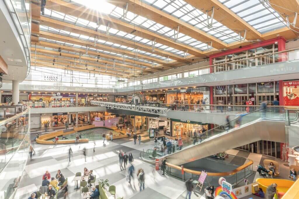 Modernes Einkaufszentrum mit Geschäften, Kunden und Tageslicht durch Glasdach. Lebendige Szene mit Rolltreppen.