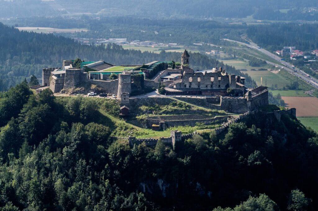 Luftaufnahme einer mittelalterlichen Burg auf einem Hügel, umgeben von Wald mit Talblick im Hintergrund.