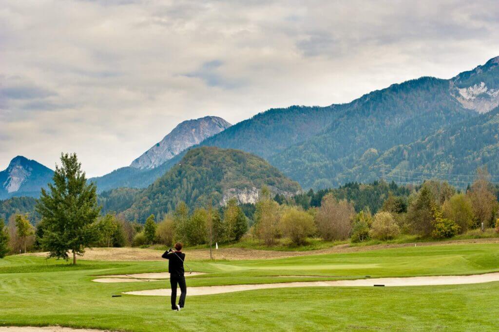 Golfer auf einem grünen Platz vor malerischer Berglandschaft, beim Abschlag in einer alpinen Umgebung.