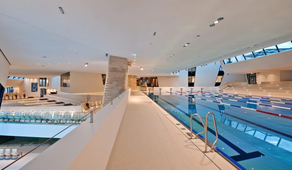 Modernes Hallenbad mit Bahnen, heller Innenraum, Architektur und Sitzbereich, ideal für Sport und Entspannung.
