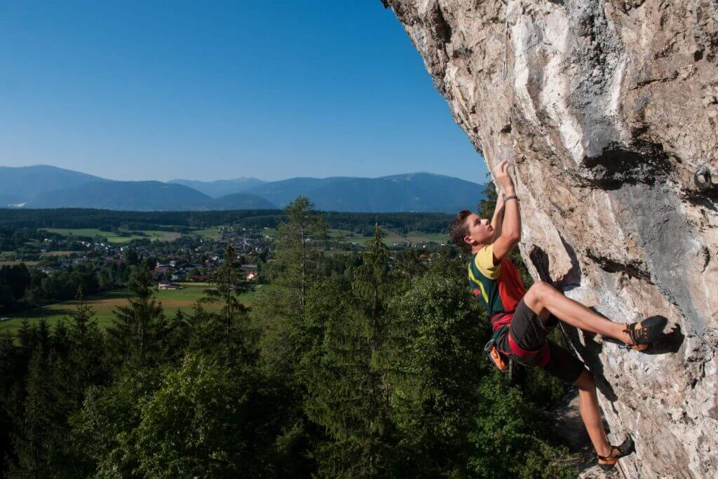 Kletterer an einer Felswand mit malerischem Blick auf ein Tal und Berge unter klarem Himmel. Abenteuer und Outdoor-Erlebnis.