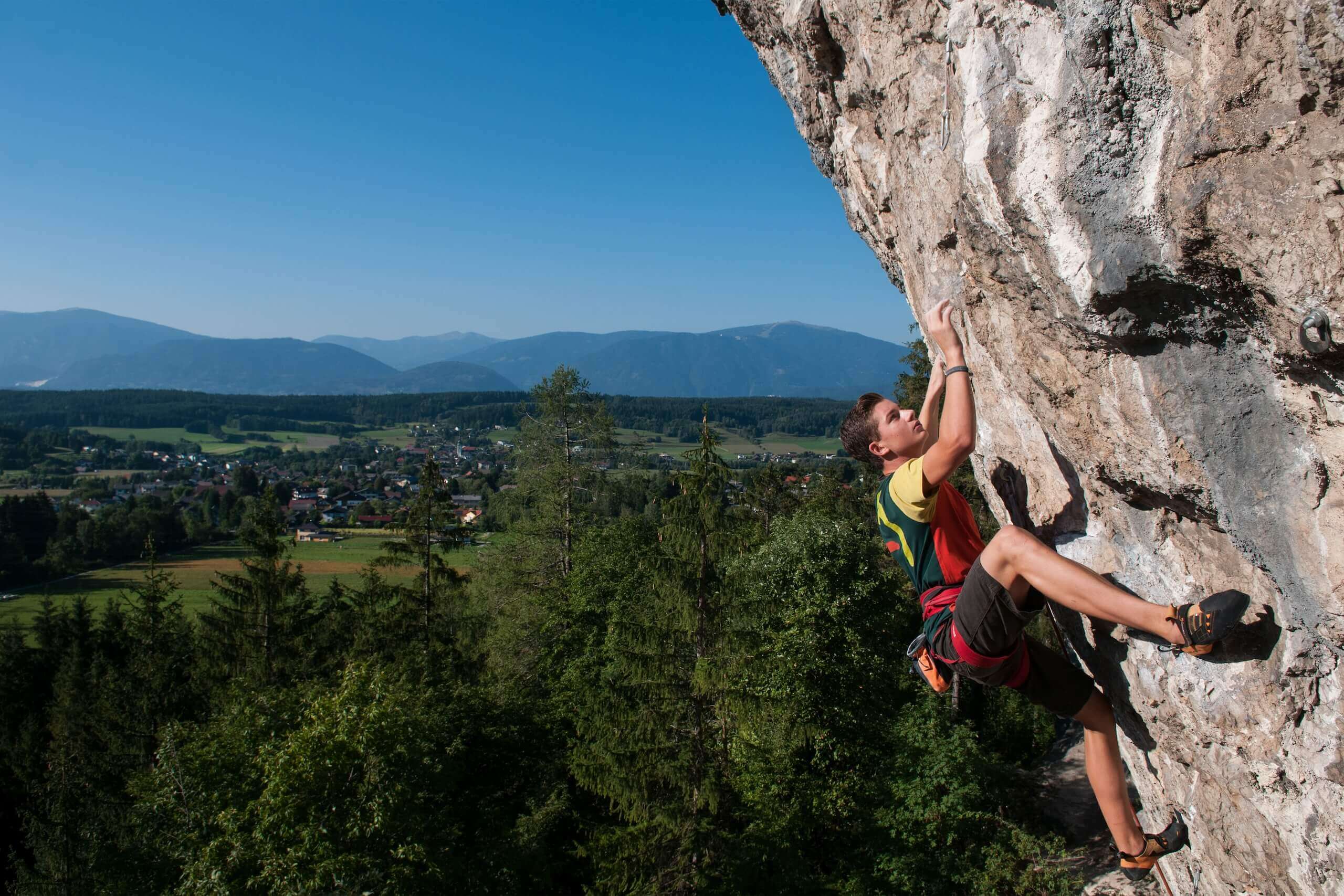 Kletterer an einer Felswand mit malerischem Blick auf ein Tal und Berge unter klarem Himmel. Abenteuer und Outdoor-Erlebnis.