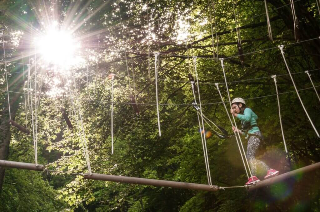 Kind im Kletterpark balanciert auf Seilbahn im Sonnenlicht, mit Helm und Sicherheitsausrüstung. Abenteuer im Wald.