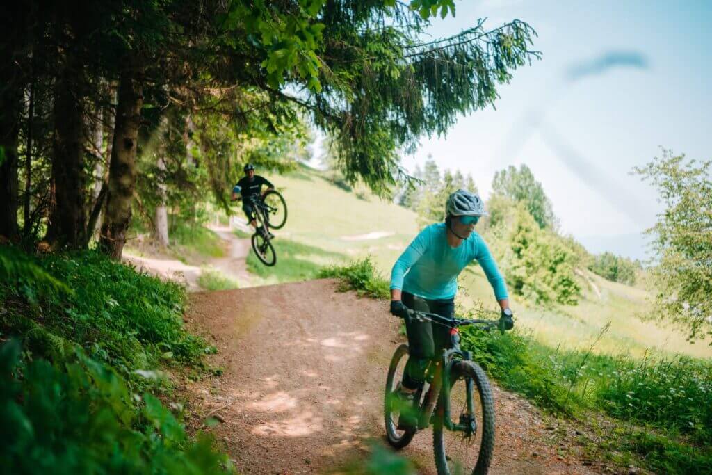 Zwei Mountainbiker fahren auf einem Waldweg; einer springt in die Luft, der andere fährt voraus.