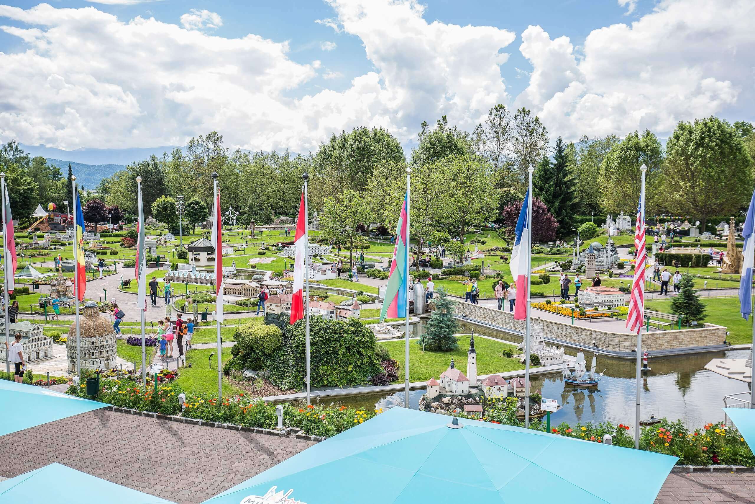 Miniaturpark mit internationalen Flaggen und Besuchern bei sonnigem Wetter, eingebettet in grüne Landschaft.