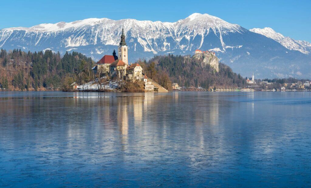 Winterliches Panorama des Bleder Sees mit schneebedeckten Bergen und der Inselkirche in Slowenien.