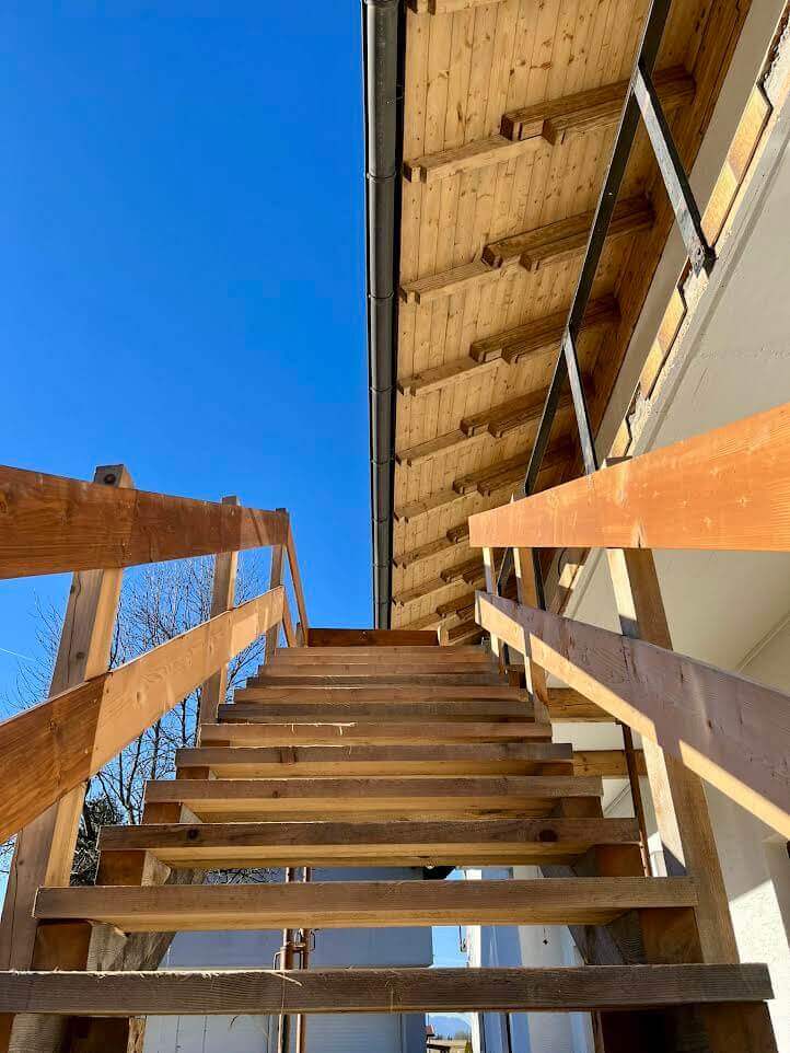 Holztreppe führt unter strahlend blauem Himmel zu einem Gebäude mit Holzbalkon. Nachhaltiges Bauen.
