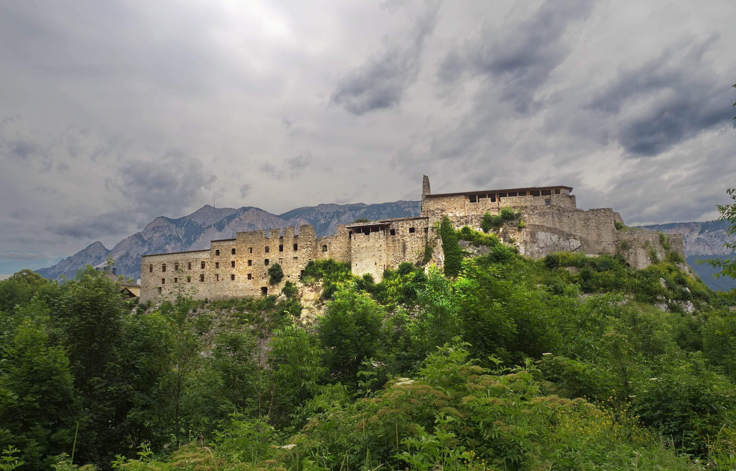 Alte Steinburg vor Bergkulisse mit bewölktem Himmel und üppigem Grün im Vordergrund.
