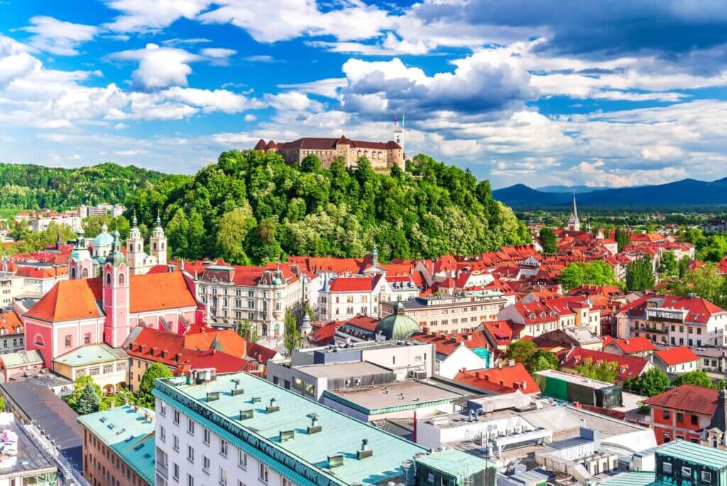 Blick auf die Altstadt von Ljubljana mit der Burg auf einem bewaldeten Hügel unter blauem Himmel und Wolken.