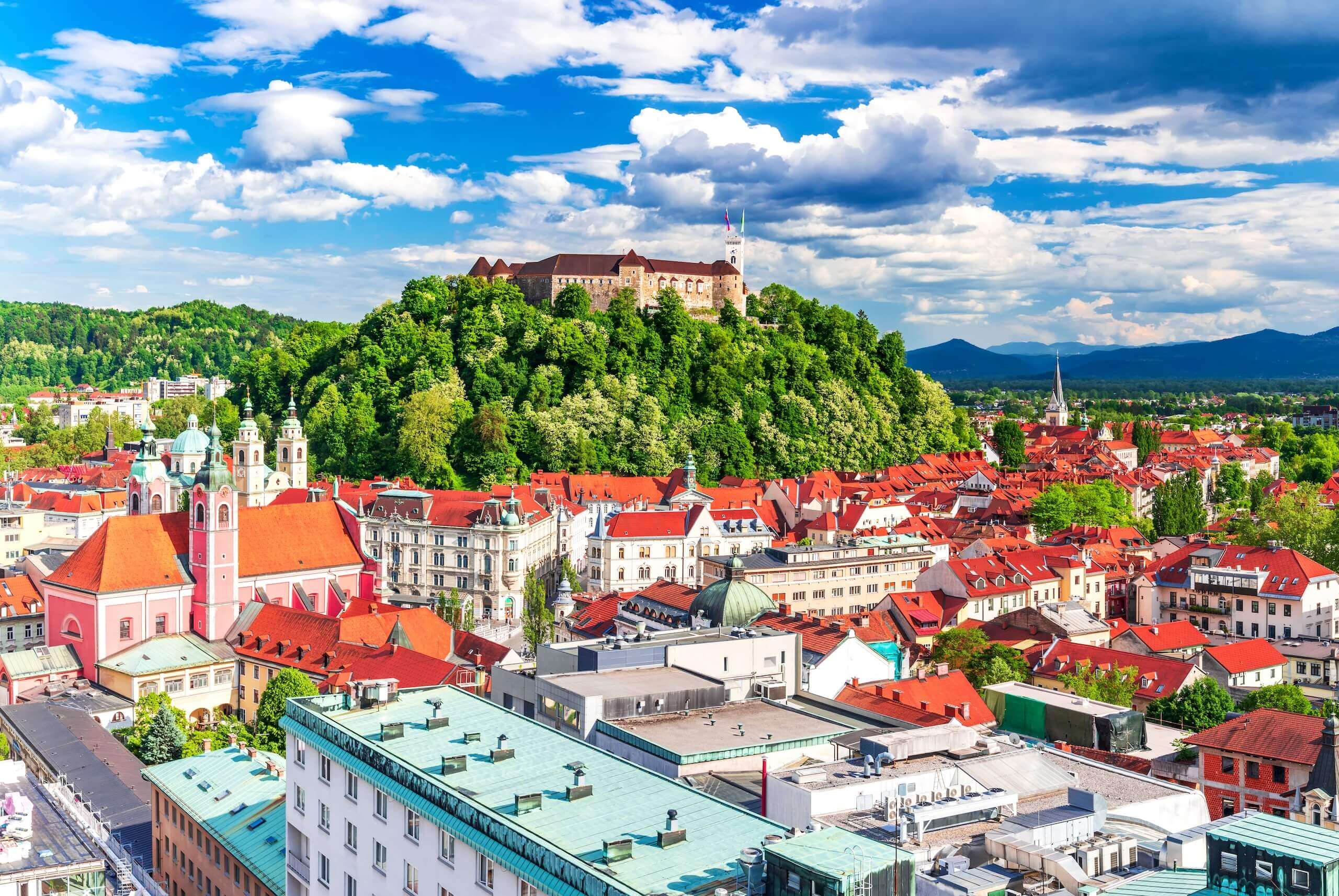 Blick auf die Altstadt von Ljubljana mit der Burg auf einem bewaldeten Hügel unter blauem Himmel und Wolken.