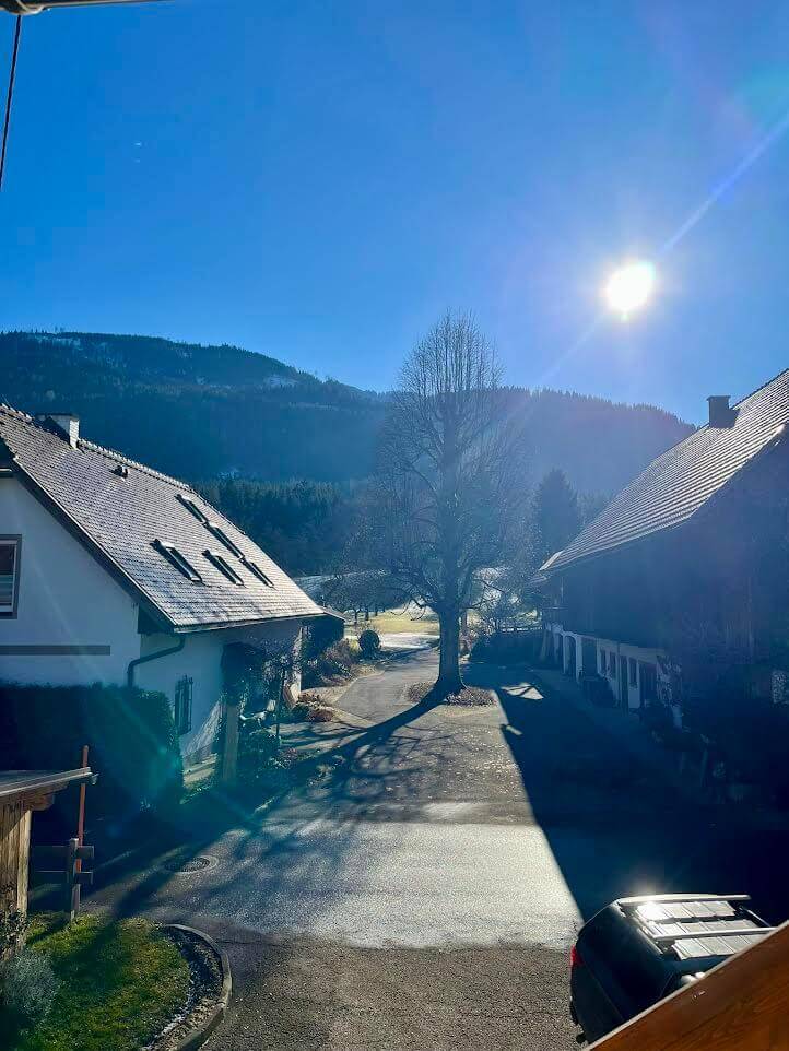 Dorfstraße bei klarem Himmel und Sonnenschein, umgeben von traditionellen Häusern und Bergen im Hintergrund.