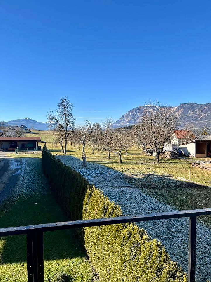 Blick auf sonnige Landschaft mit Bäumen, grünen Wiesen und Bergen im Hintergrund unter klarem, blauem Himmel.