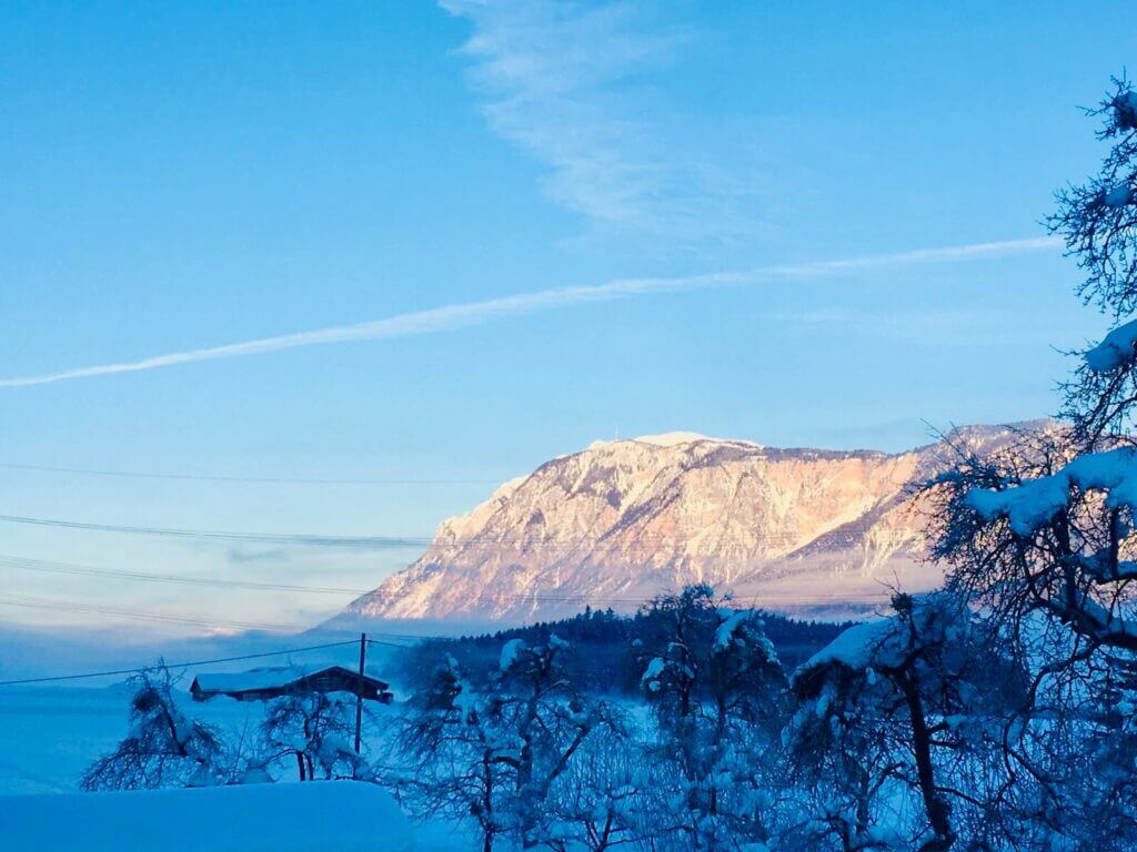Verschneite Alpen im Morgenlicht, blauer Himmel, schneebedeckte Bäume und eine Hütte im Vordergrund.
