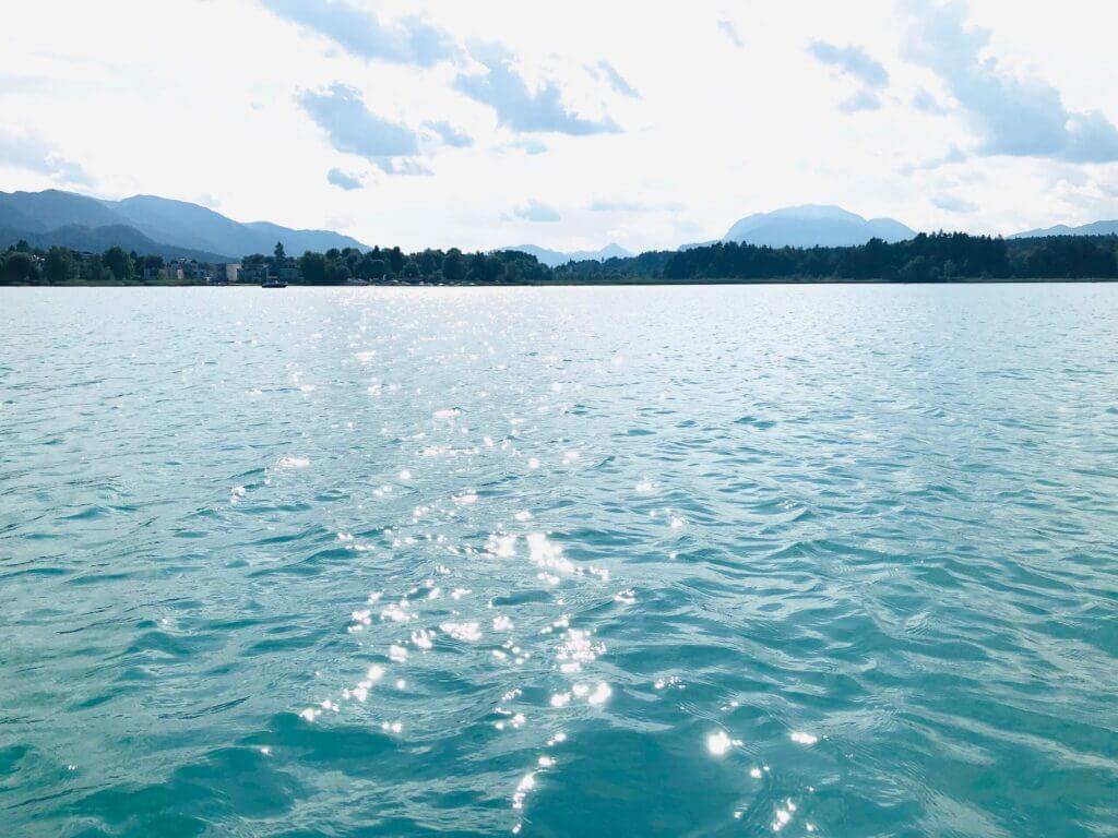 Blick auf funkelnde Wasseroberfläche eines Sees bei Sonnenschein, umgeben von Bergen und bewölktem Himmel.