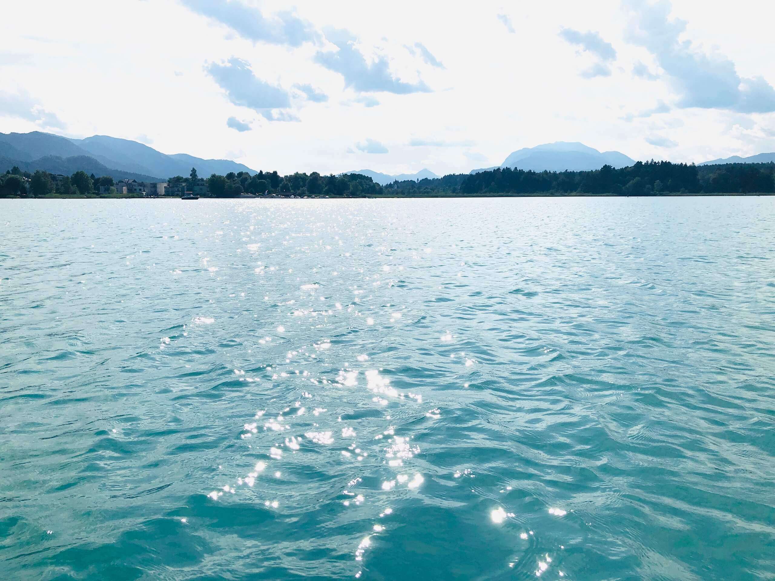 Blick auf funkelnde Wasseroberfläche eines Sees bei Sonnenschein, umgeben von Bergen und bewölktem Himmel.