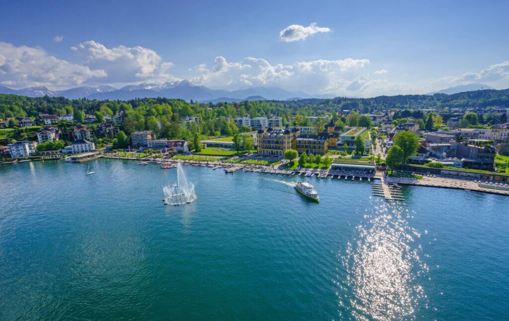 Blick auf den Wörthersee mit Promenade, segelndem Boot und Bergen im Hintergrund an einem sonnigen Tag.