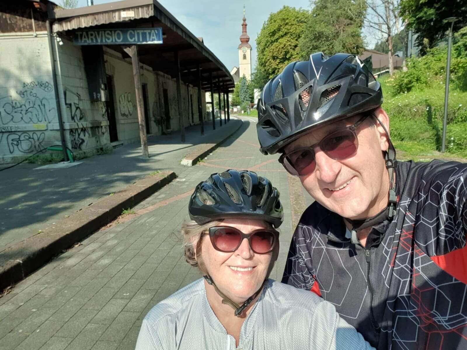 Paar mit Fahrradhelmen vor dem alten Bahnhof Tarvisio Città, sonniger Tag, Kirche im Hintergrund.