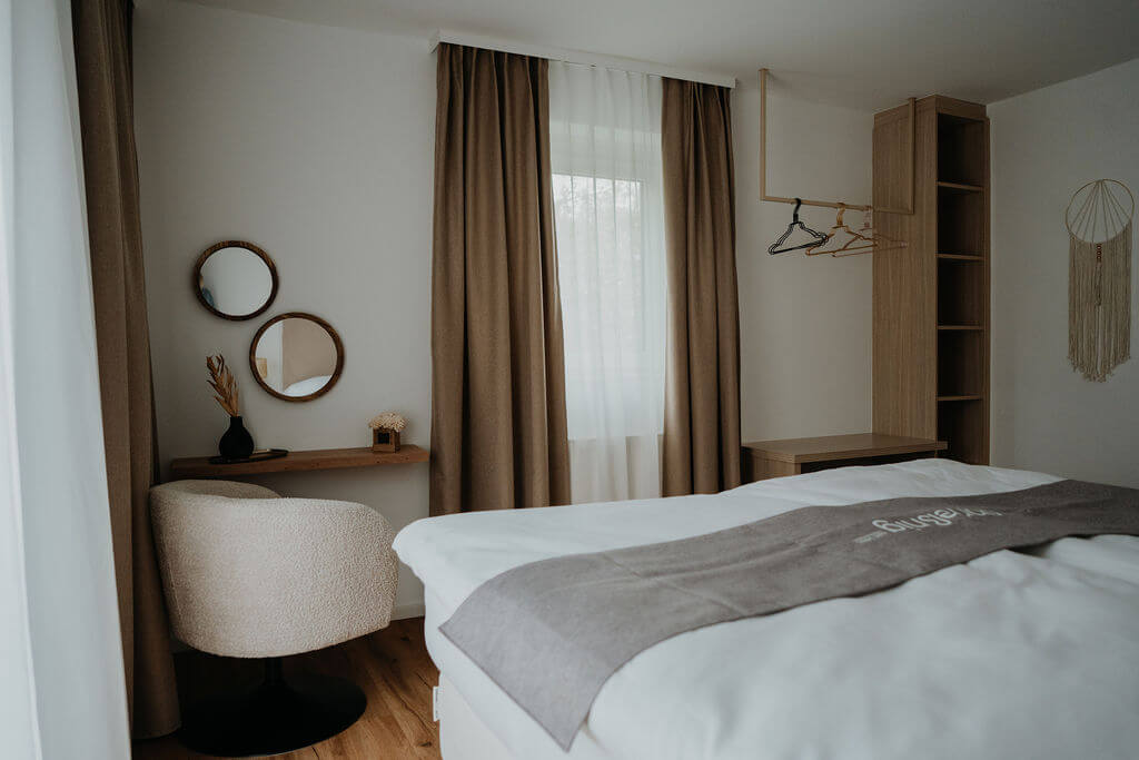 Modernes, gemütliches Schlafzimmer mit beigem Sessel, Holzmöbeln, Spiegeln und Dekor vor Fenster mit Vorhang.
