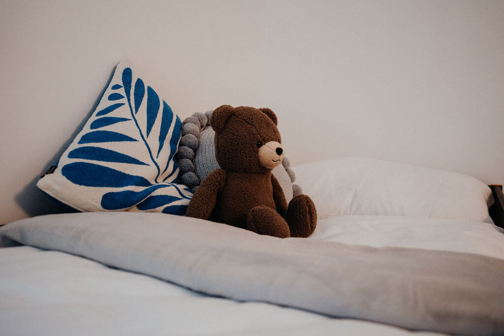 Braunbär-Plüschspielzeug auf Bett mit blauem Kissen; gemütliches Kinderzimmer-Setup.