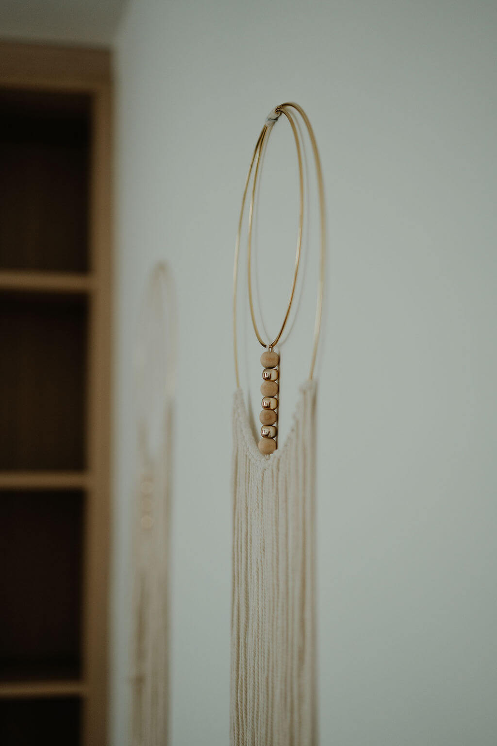 Minimalistisches Makramee-Wanddekor mit Holzperlen und goldenen Ringen, vor einem neutralen Hintergrund.