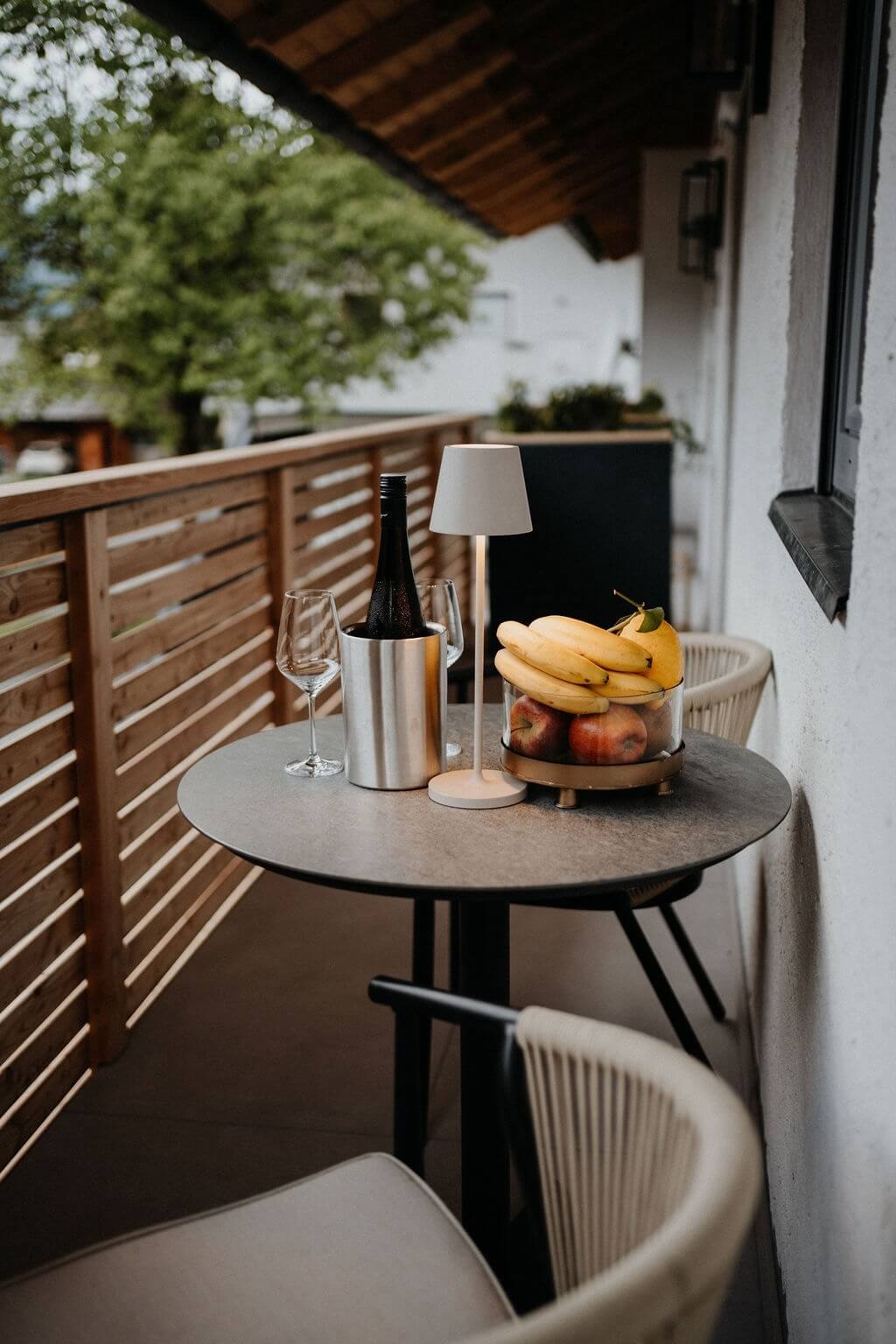 Weinflasche, Obstschale und Lampe auf einem Balkon-Tisch mit Aussicht auf Natur und Ruhe.