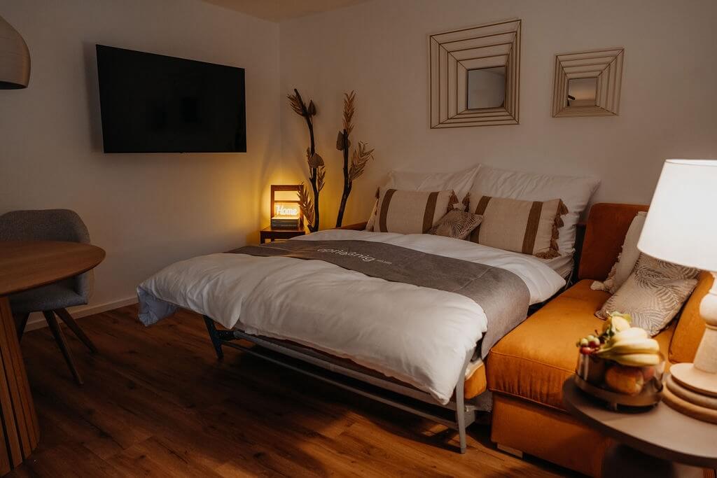 Gemütliches Schlafzimmer mit Schlafsofa, Fernseher, dekorativen Kissen und stimmungsvollem Licht auf Holzfußboden.