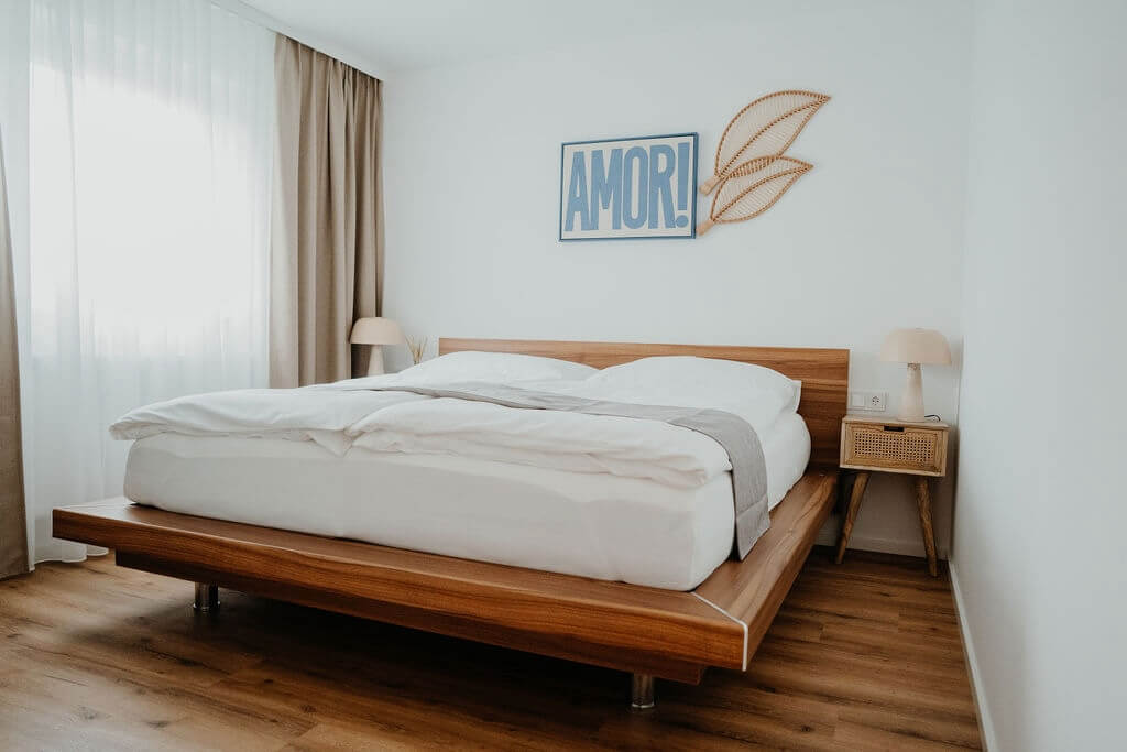 Modernes Schlafzimmer mit Holzbett, weißer Bettwäsche und dekorativer AMOR!-Wandkunst, gemütlich und stilvoll gestaltet.