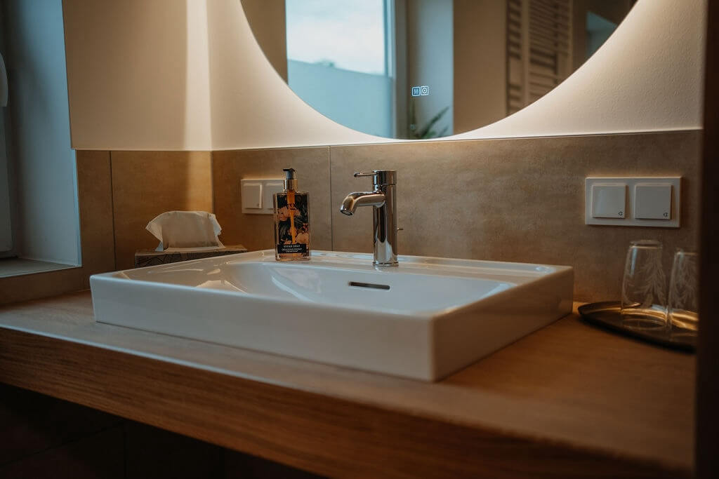 Modernes Badezimmer mit Wasserhahn, Waschbecken und Spiegel im stimmungsvollen Licht, inklusive Seifenflasche und Handtüchern.