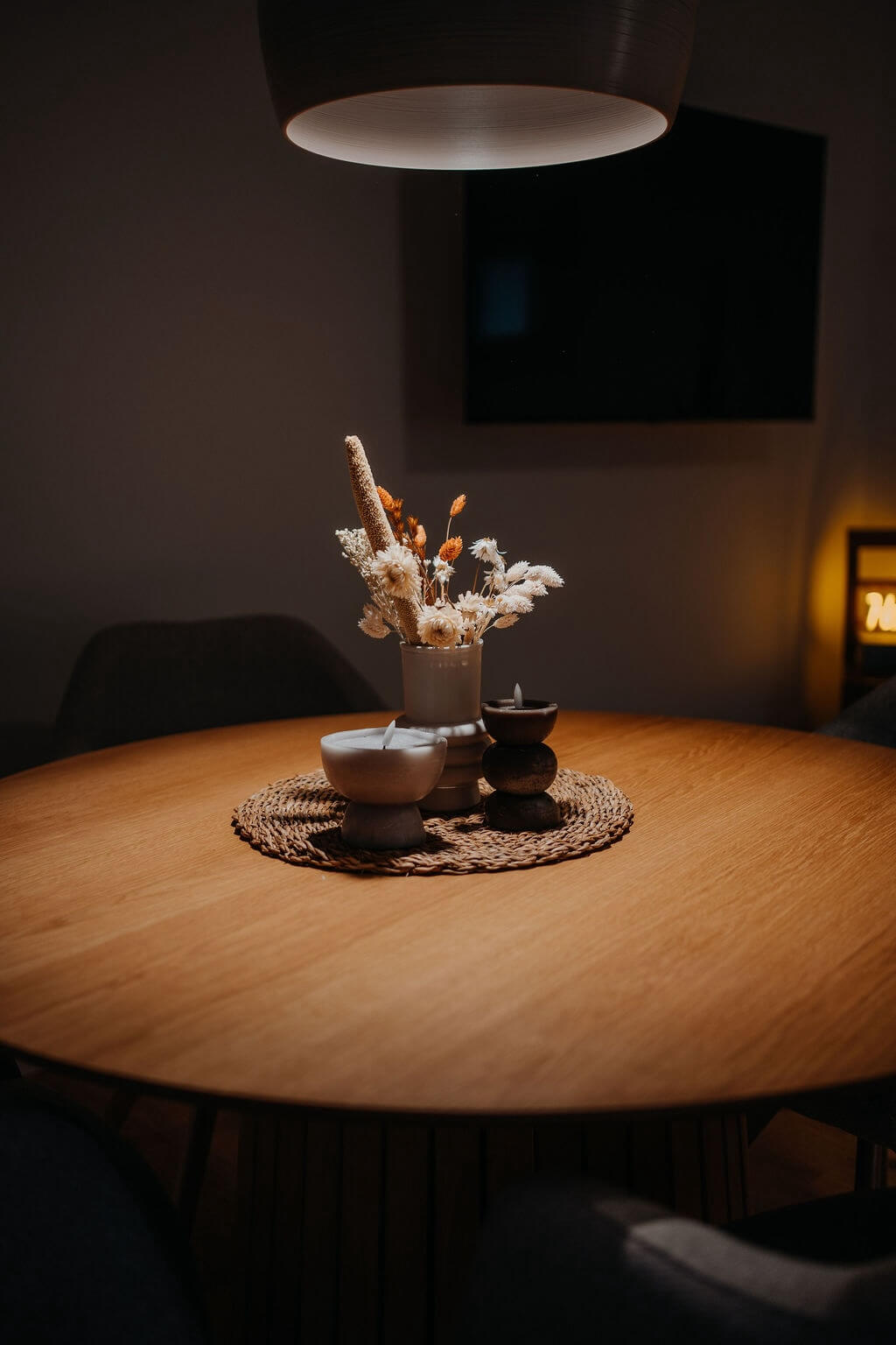 Runder Holztisch mit Trockenblumen in Keramikvase und Kerzen, in sanft beleuchtetem Raum dekoriert.