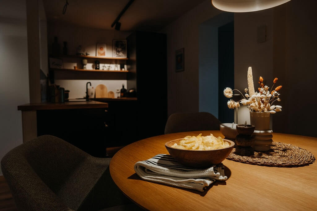 Gemütlicher Essbereich mit Holzdeko und Snacks, im Hintergrund eine stilvolle, dunkle Küche.